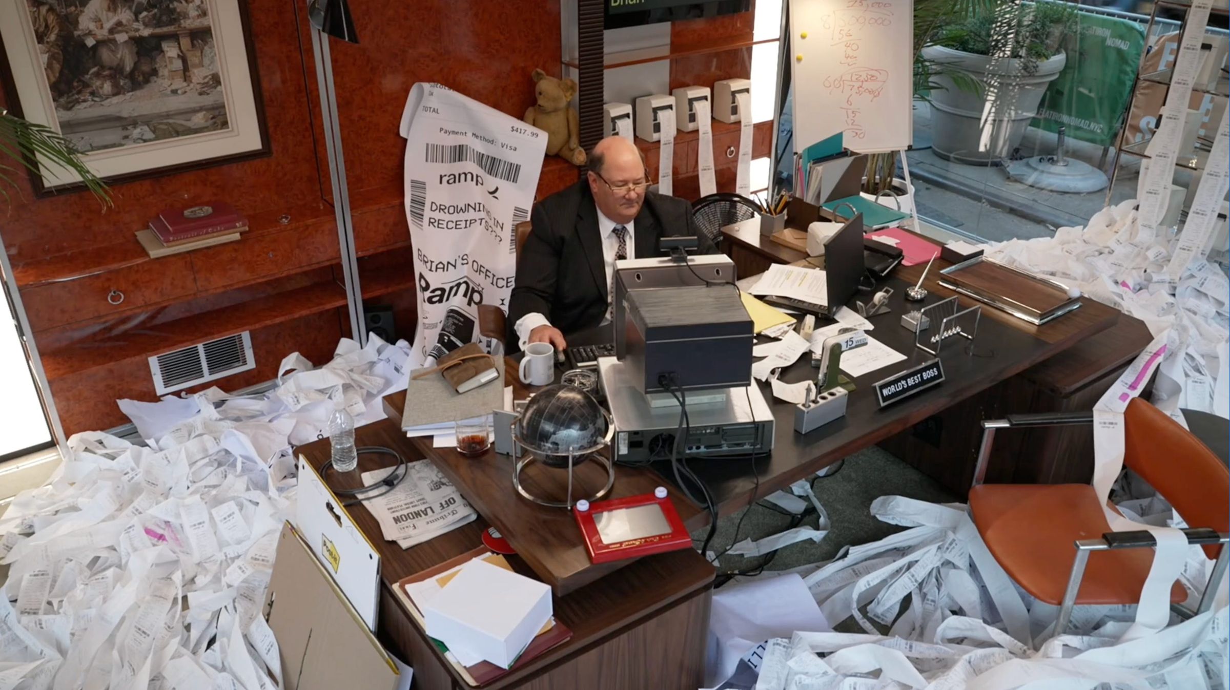 Brian Baumgartner at a desk buried in receipt paper, receipt printers on the wall behind him
